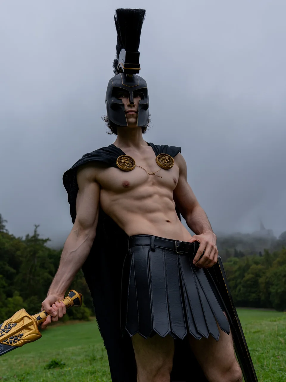 Agamemnon cosplay from The Odyssey (2026) in black-and-gold armor with a crested helmet and sword in a misty field.