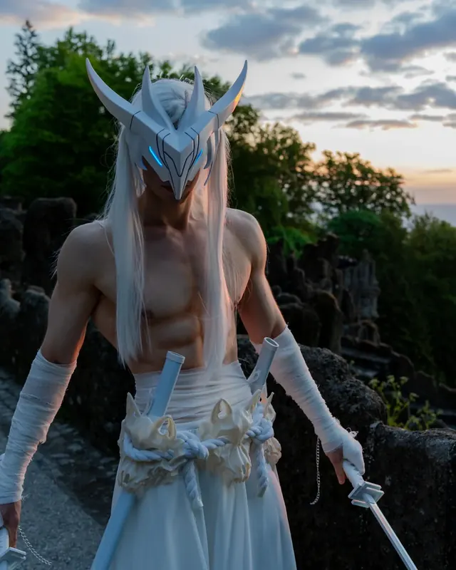 White fantasy cosplay with a sculpted mask, long wig and prop weapons at sunset.