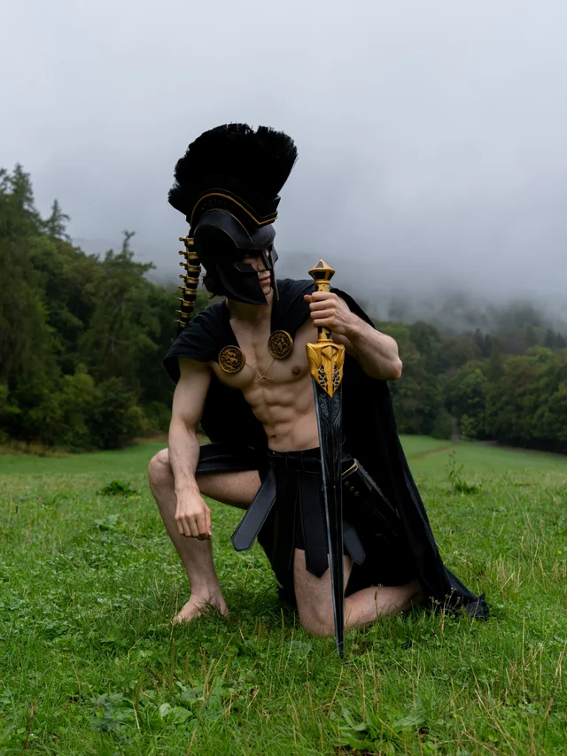 Agamemnon cosplay kneeling with a sword in a misty green landscape.