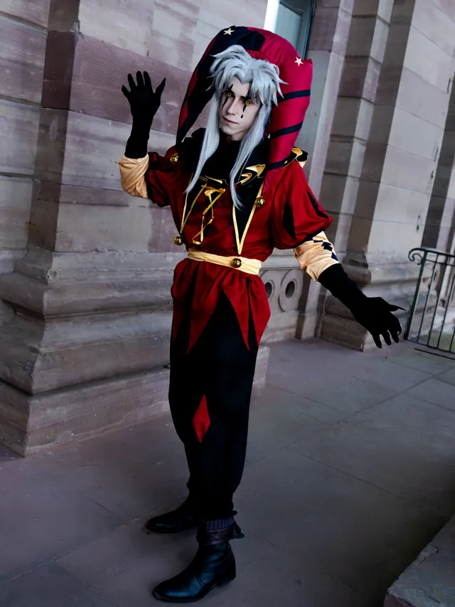 Full-body Pierrot cosplay in a red and black jester costume beside a stone column.
